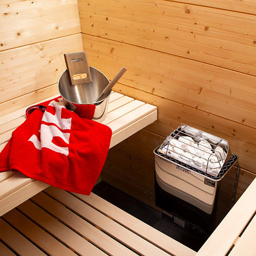 Harvia KIP W-series stainless-steel heater beside a bench with a bucket, ladle, and red towel.